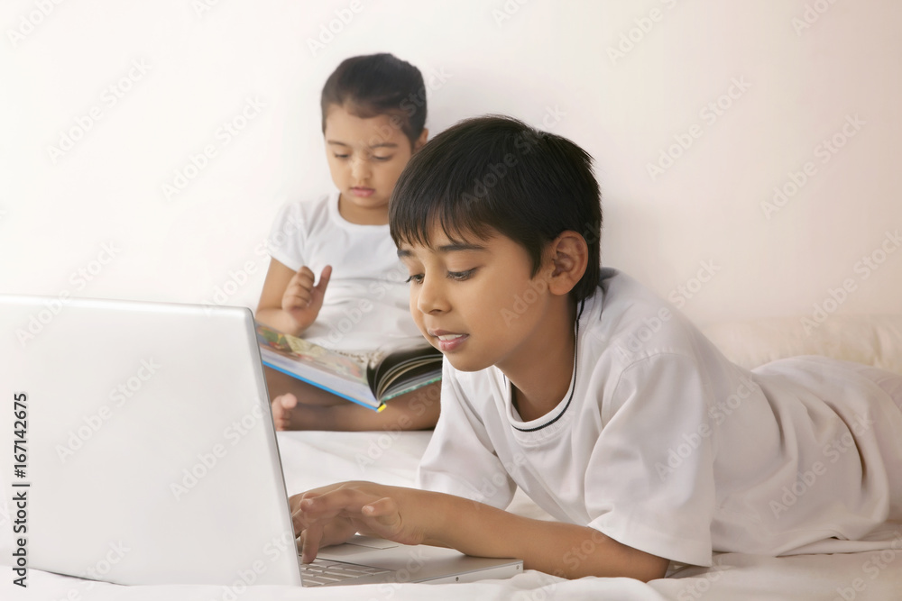 Boy using laptop while girl reading book in bed Boy using laptop while girl reading book in bed