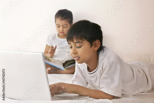 Boy using laptop while girl reading book in bed