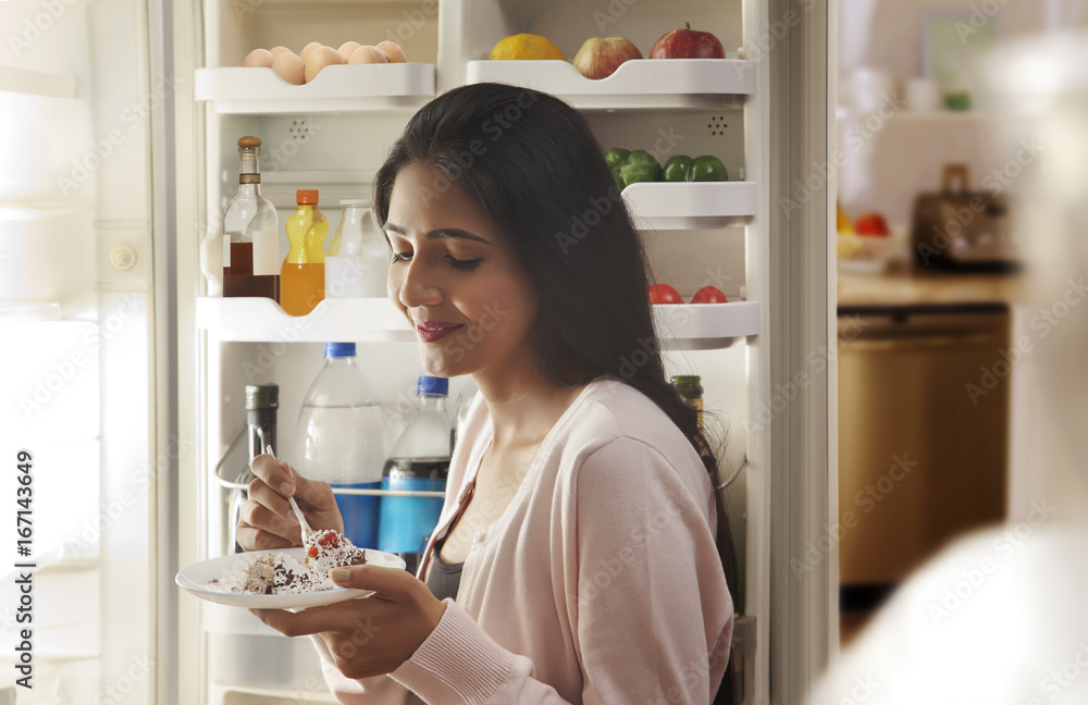 Young woman eating cake in front of open refrigerator 