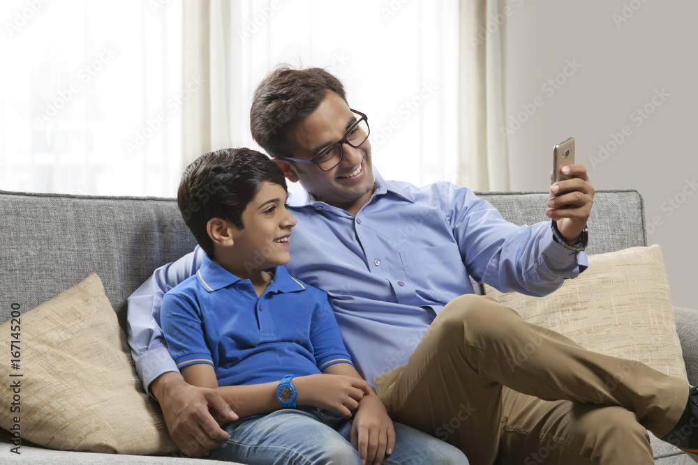 Smiling father and son sitting on sofa looking at mobile phone Smiling father and son sitting on sofa looking at mobile phone