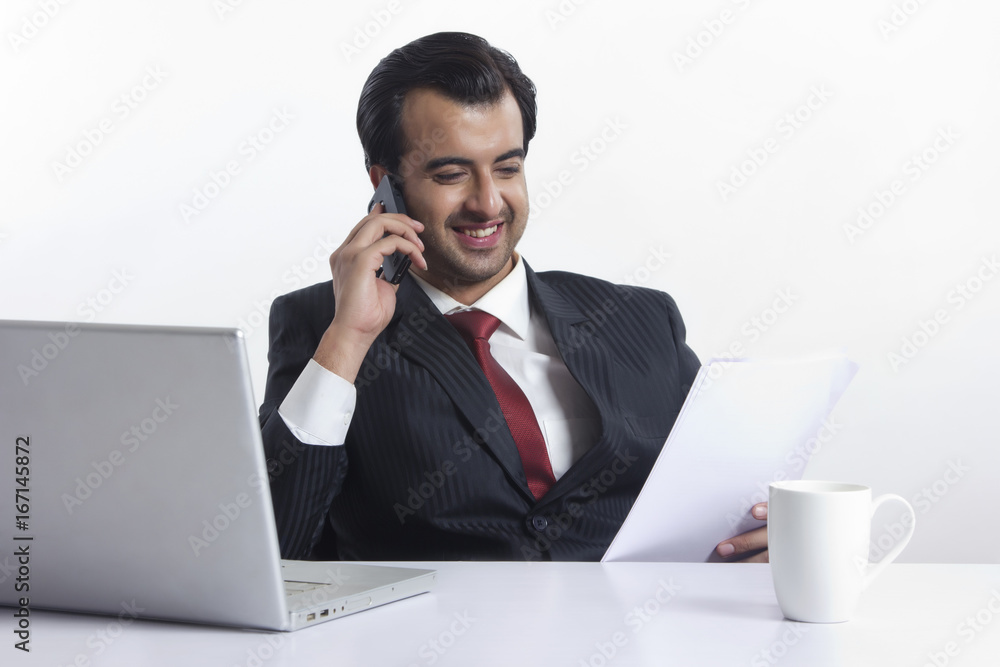 Happy businessman reading document while answering smart phone at desk over white background Happy businessman reading document while answering smart phone at desk over white background