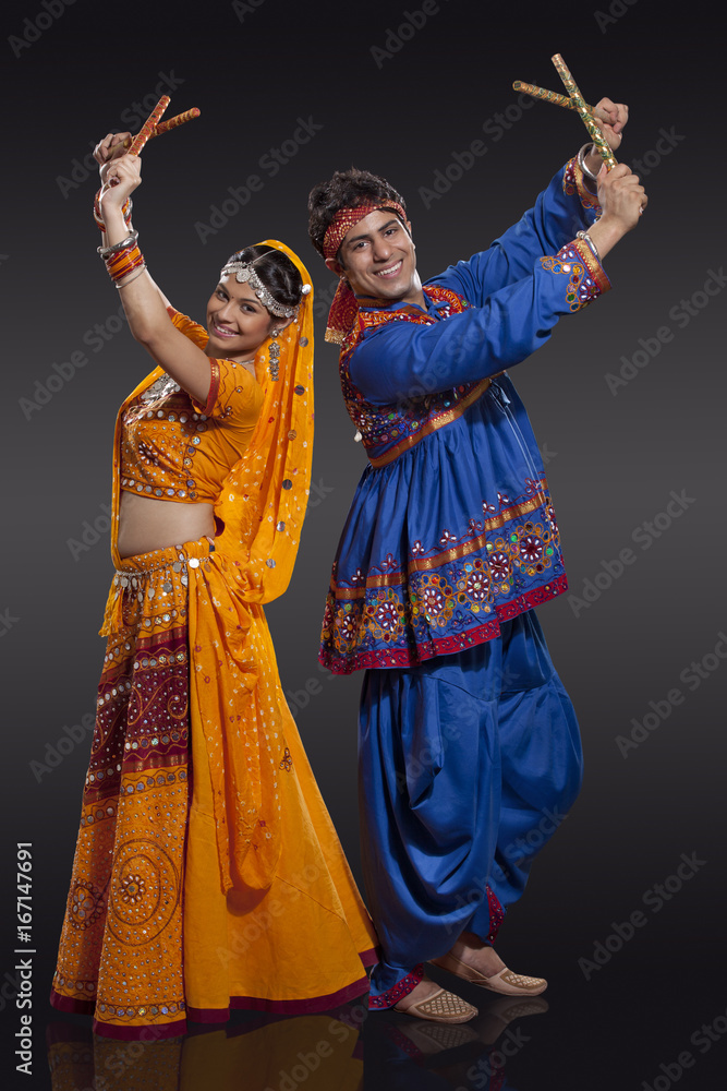 Full length portrait of happy young Indian couple performing Dandiya Raas over black background