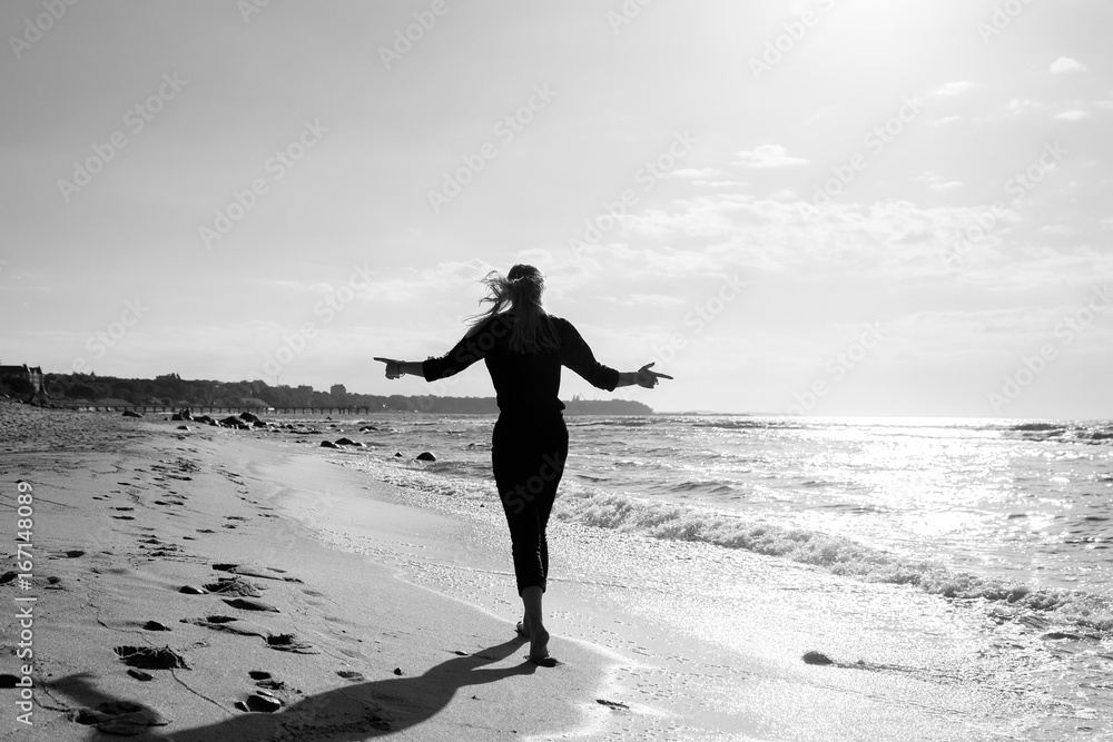 sad blond barefoot woman walking away at sunny sea beach, monochrome image