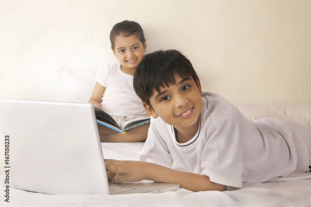 Portrait of boy using laptop with girl reading book in bed Portrait of boy using laptop with girl reading book in bed