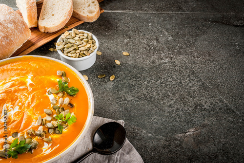 Traditional fall and winter dishes, hot and spicy pumpkin  soup with pumpkin seeds, cream and freshly baked baguette, on black stone table, copy space