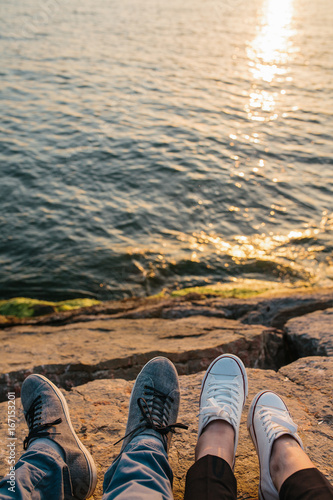 Male and female feet in sneakers on the rocky shore with beautiful view on wavy water, friendship or love concept. Trust and travel together.