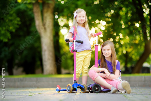 Foto Small children learning to ride scooters in a city park on sunny summer evening