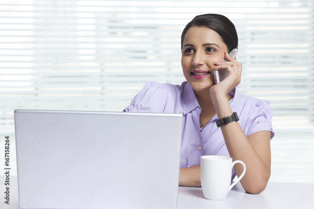Smiling businesswoman answering smart phone at office desk Smiling businesswoman answering smart phone at office desk