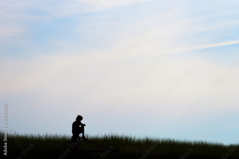 Silhouette of young teen man, curly hair, sitting on post in field, deep in thought beneath a pastel morning or evening sky