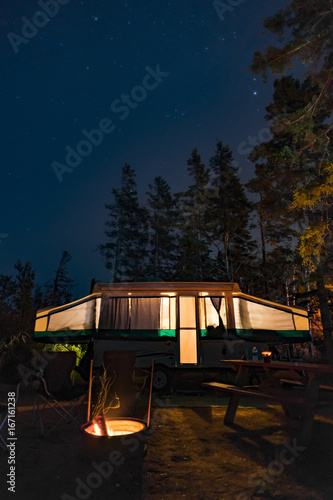 Campsite at night with glow from RV trailer and campfire, tall pine trees and night stars