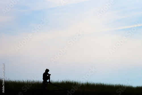 Silhouette of young teen man, curly hair, sitting on post in field, deep in thought beneath a pastel morning or evening sky