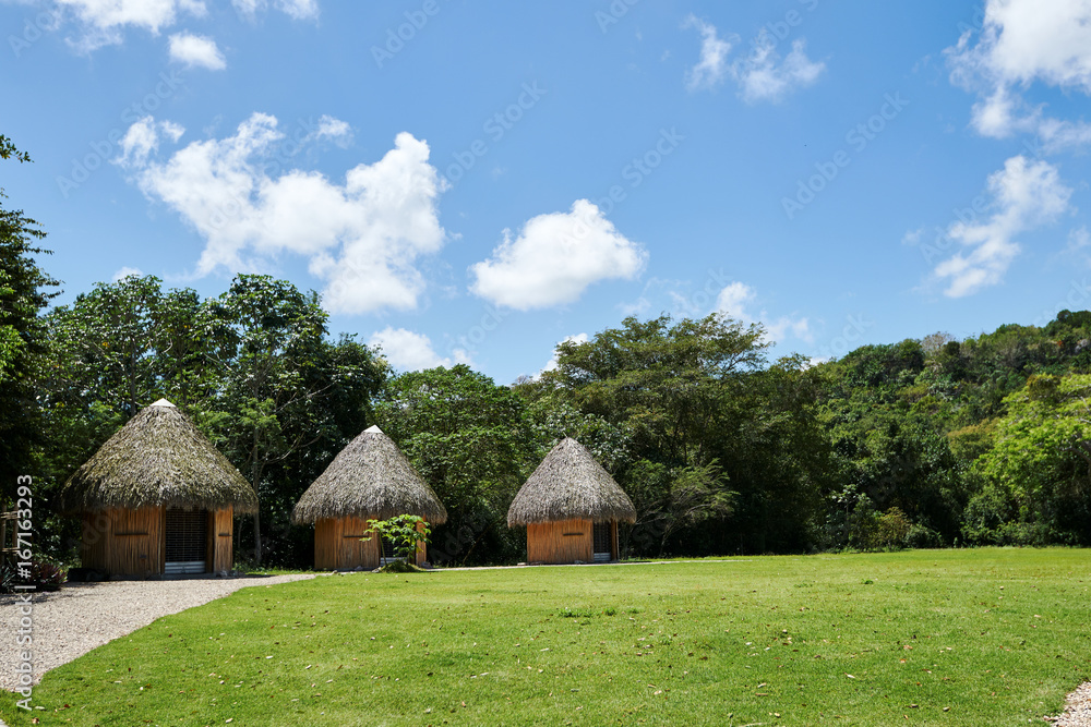 Tropical huts on an island with conical thatch roofs, weaved out of coconut palm tree fronds. Bright daylight, blue sky with puffy Cumulus clouds. Copy space. Tropical huts on an island with conical thatch roofs, weaved out of coconut palm tree fronds. Bright daylight, blue sky with puffy Cumulus clouds. Copy space.
