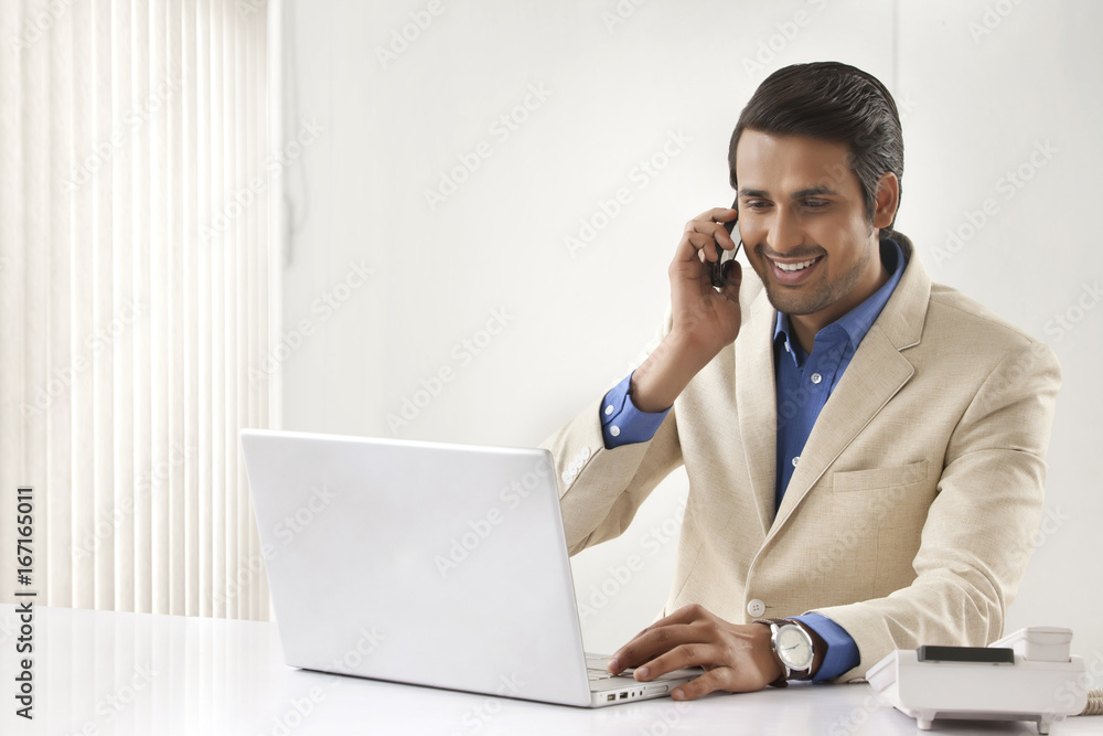 Handsome young businessman on call using laptop at office desk Handsome young businessman on call using laptop at office desk