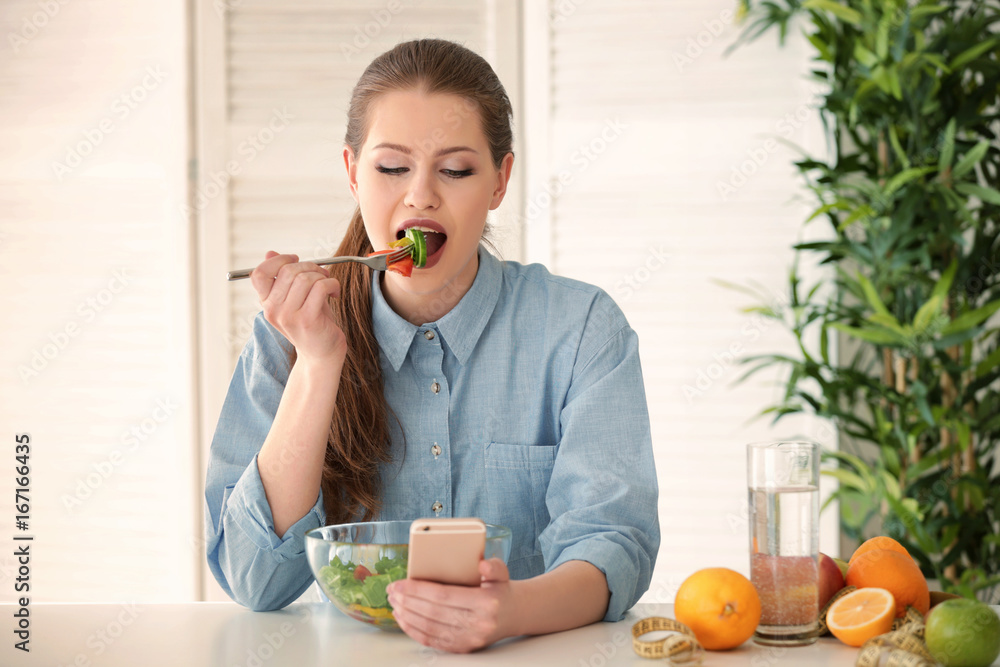 Young woman using mobile phone for counting calories while eating salad. Weight loss concept Young woman using mobile phone for counting calories while eating salad. Weight loss concept