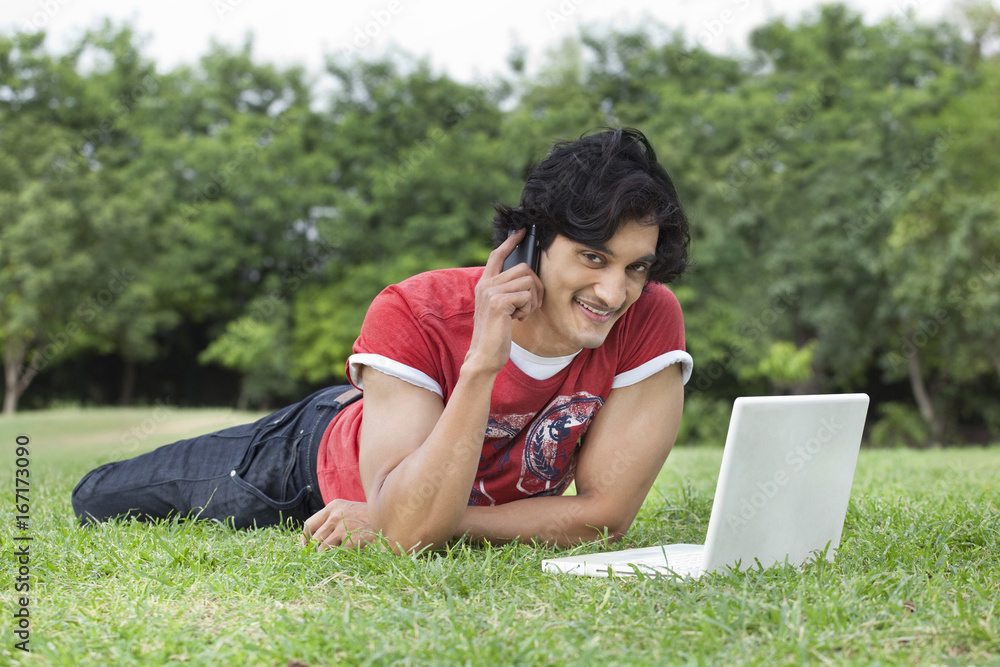 Portrait of smiling young man using cell phone and laptop while lying on grass Portrait of smiling young man using cell phone and laptop while lying on grass