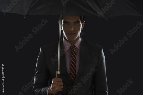 Portrait of confident young businessman holding umbrella against black background