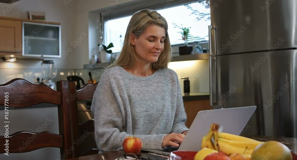 A woman working at home on her computer takes a break and bites into an apple. She looks at the camera happily.