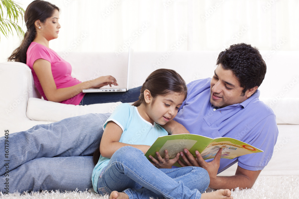 Smiling father helping child in homework with woman using laptop in the background 