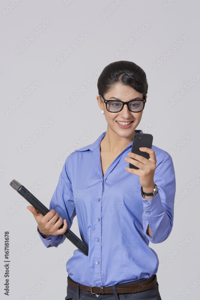 Happy businesswoman using mobile phone while holding laptop against gray background Happy businesswoman using mobile phone while holding laptop against gray background