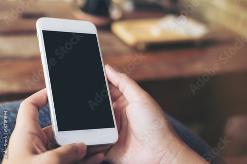 Mockup image of hands holding white mobile phone with blank black screen with blur wooden table background in cafe