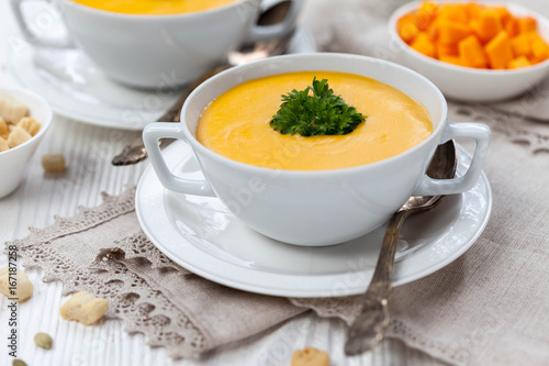 Pumpkin cream soup with croutons, raw fresh pumpkin pieces  and herbs on a white rustic wooden background