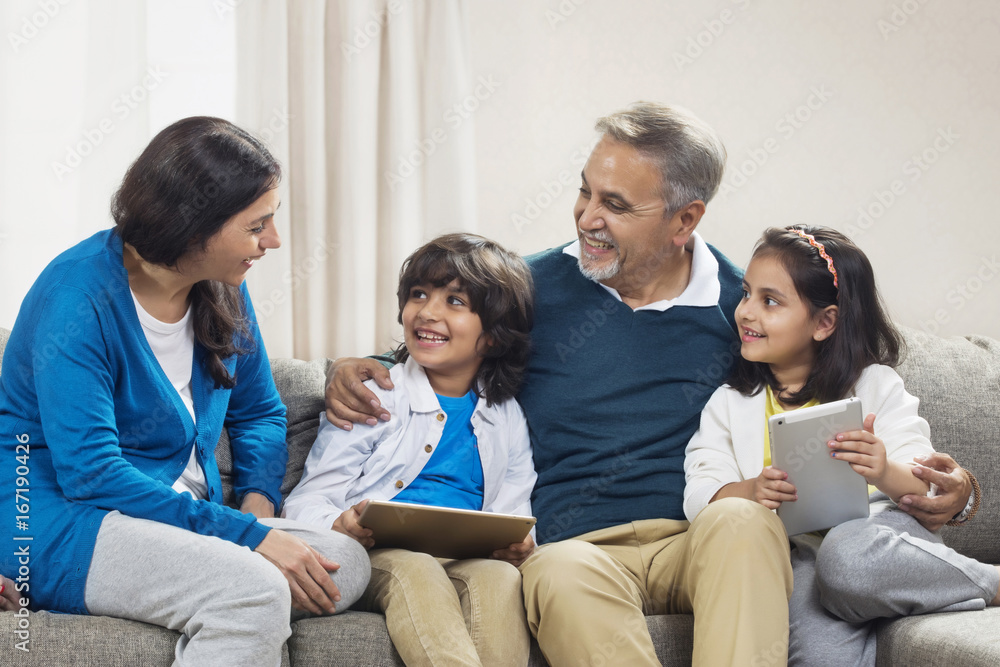 Smiling grandchildren using digital tablet with grandparents sitting on sofa Smiling grandchildren using digital tablet with grandparents sitting on sofa