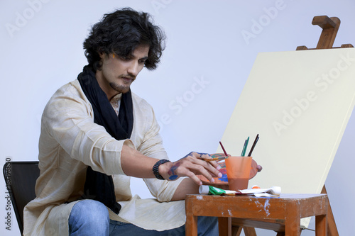 Young artist sitting near easel against colored background 