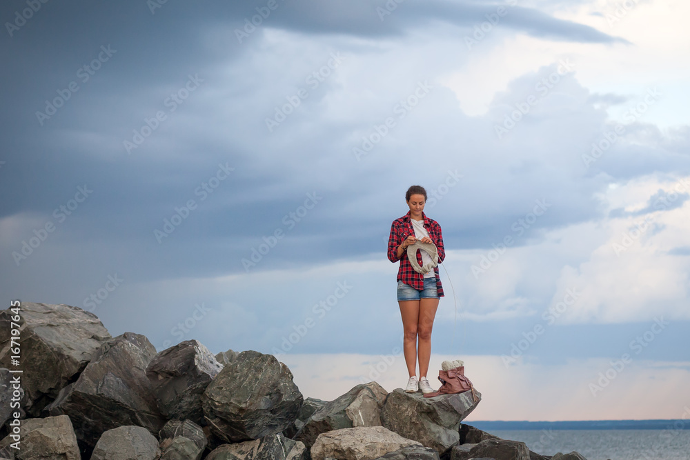 A young d woman in a checkered red shirt, white T-shirt and jeans shorts knits a gray sweater from natural woolen threads and stands on the rocks near the sea before the rain, the sky in the clouds