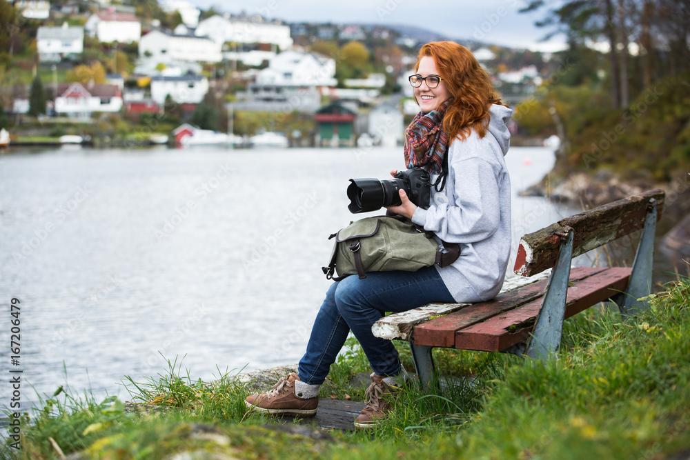 Fototapeta premium Girl photographer with red hair sitting on a wooden bench in Scandinavia