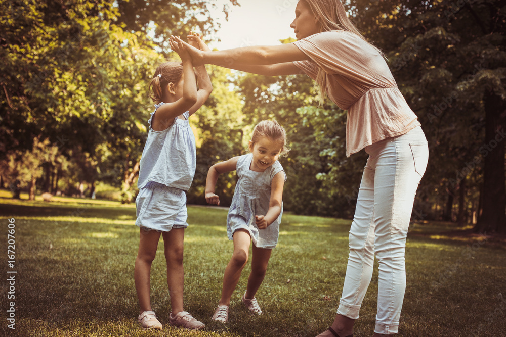 Fototapeta premium Mother with two child in meadow. Mother holding hands with girl other girls passing under the arm