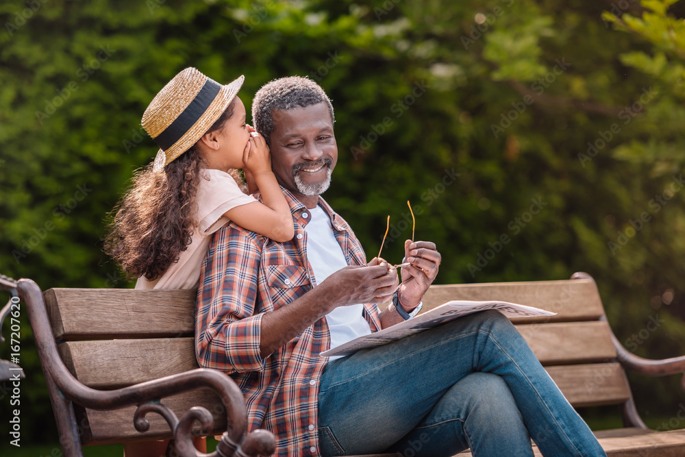 little adorable african american grandchild whispering to her smiling grandfather while sitting on bench in park - obrazy, fototapety, plakaty 