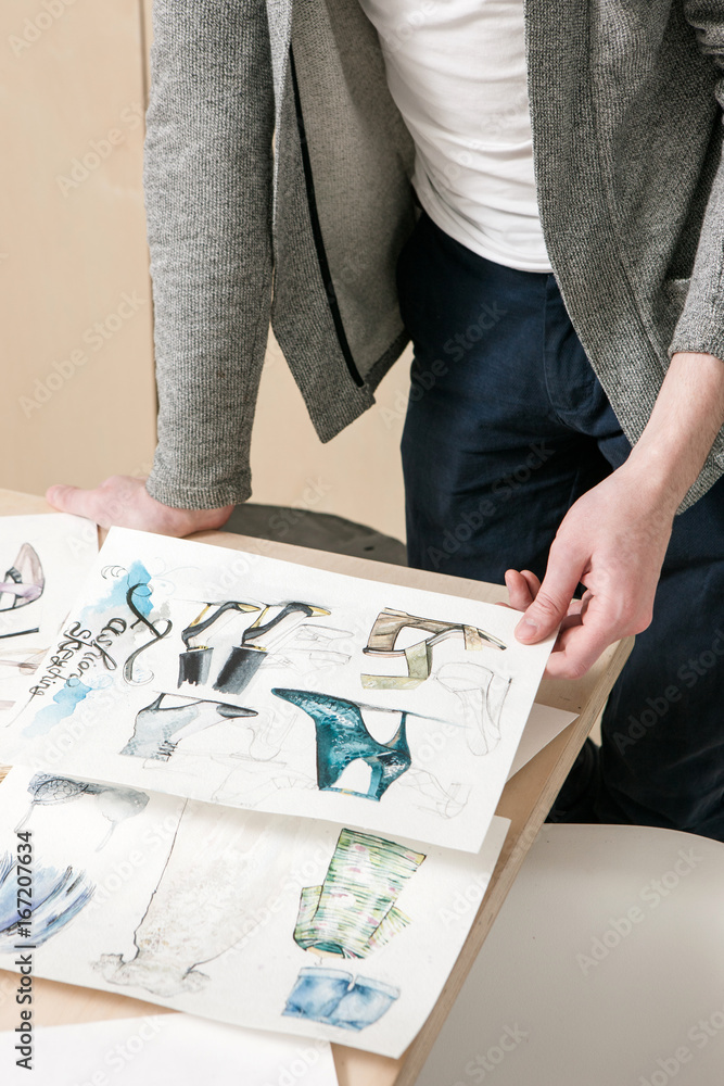 Designer work on fashion sketches. Unrecognizable man with pictures of clothes and footwear in hands. New collection, vogue project, creative artist exam concept Designer work on fashion sketches. Unrecognizable man with pictures of clothes and footwear in hands. New collection, vogue project, creative artist exam concept