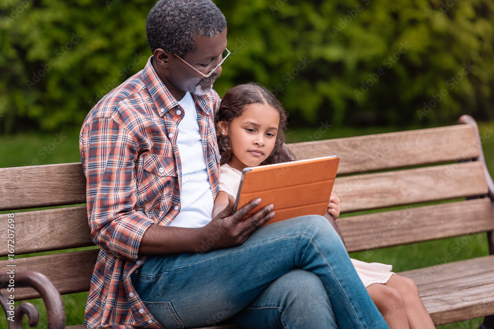adorable african american girl and her grandfather using digital tablet sitting on bench in park adorable african american girl and her grandfather using digital tablet sitting on bench in park