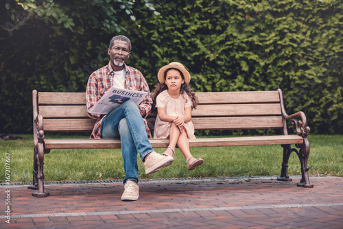 little african american granddaughter and her grandfather sitting on bench in park