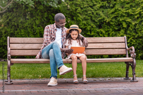 adorable african american grandchild and her grandfather listening music on digital tablet while sitting on bench in park
