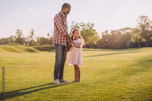african american granddaughter and her grandfather holding hands and standing on green lawn