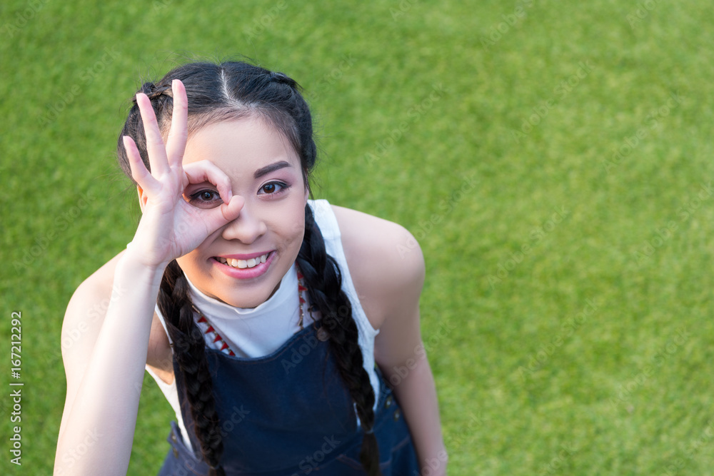 high angle view of happy young asian woman showing ok sign and smiling at camera while standing on green lawn