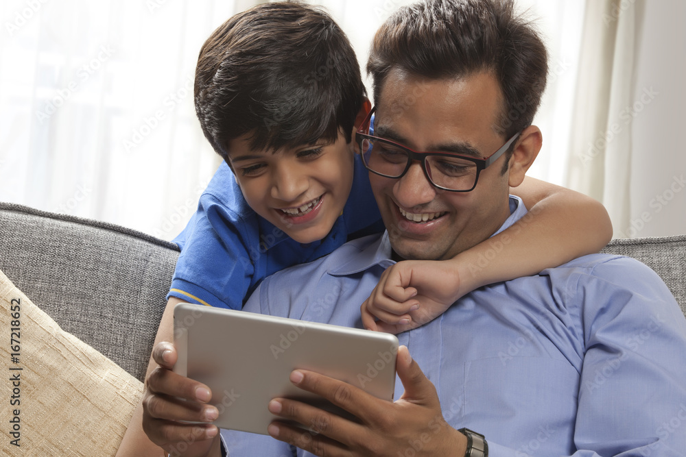 Smiling father using digital tablet sitting on sofa while his son looks from behind Smiling father using digital tablet sitting on sofa while his son looks from behind