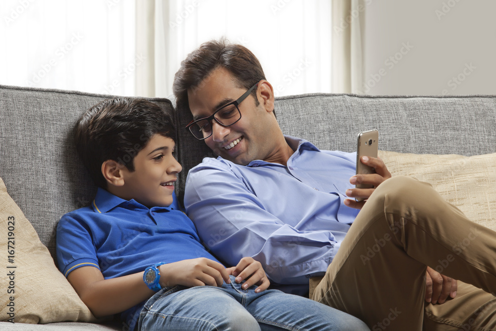 Smiling father and son sitting on sofa using mobile phone Smiling father and son sitting on sofa using mobile phone