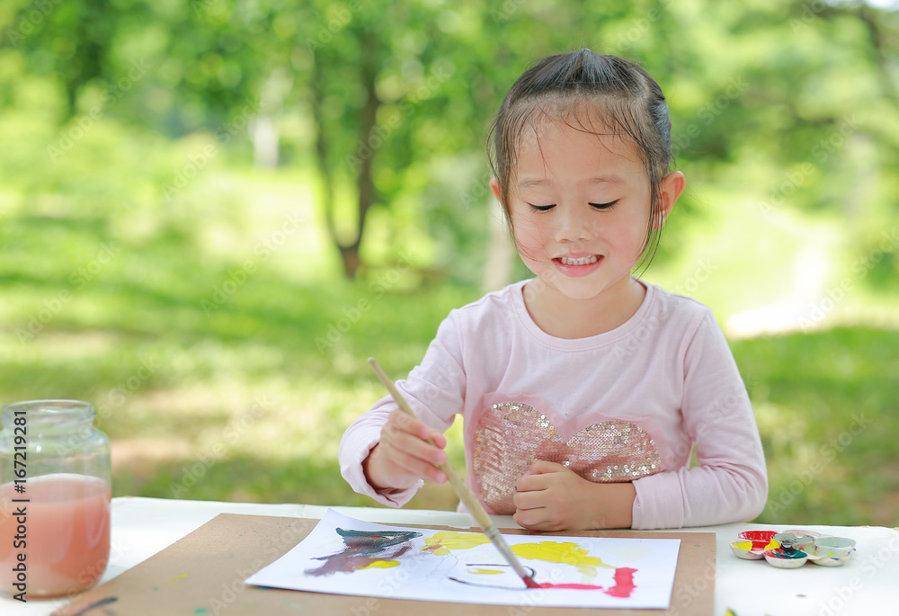 Happy asian child girl sitting at a table in a summer garden painting with paintbrush, Education art concept. Happy asian child girl sitting at a table in a summer garden painting with paintbrush, Education art concept.