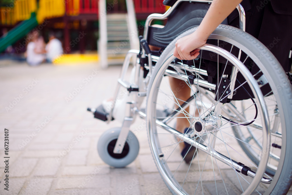 Fototapeta premium A girl with a broken leg sits in a wheelchair in front of the playground.