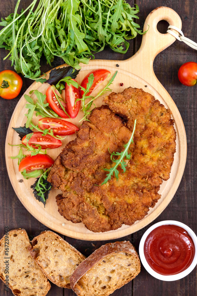 A large Viennese schnitzel and tomato salad on a cutting board on a dark wooden background. Top view. A large Viennese schnitzel and tomato salad on a cutting board on a dark wooden background. Top view.