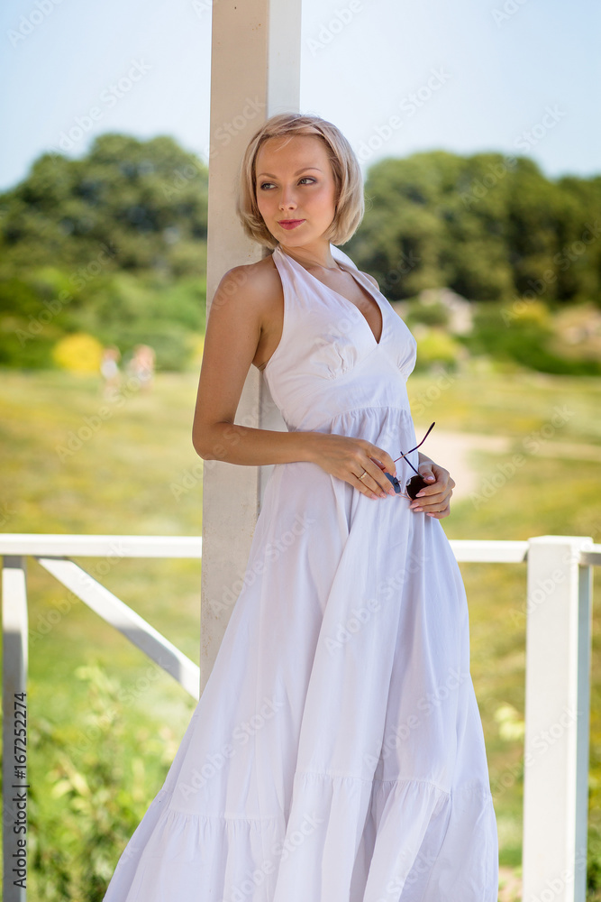 Naklejka premium Blond young female in long white dress standing in alcove. Summer day, woman posing for pictures in summer house in meadow.