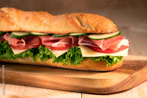 Fresh crispy baguette deli sandwich with prosciutto, cheese, tomatoes, lettuce, cucumbers on a wooden background.Close Up. Healthy concept.