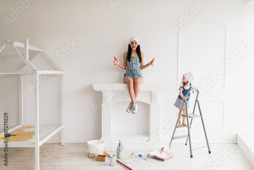 The young smiling mother does repair at home, she sits on fireplace and keeps thumbs up and her daughter stands on ladder and looks at mother