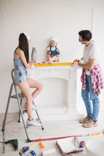 The young family does repair at home, mother and father stand and measure width of the fireplace on which their daughter sits and looks at phone photo