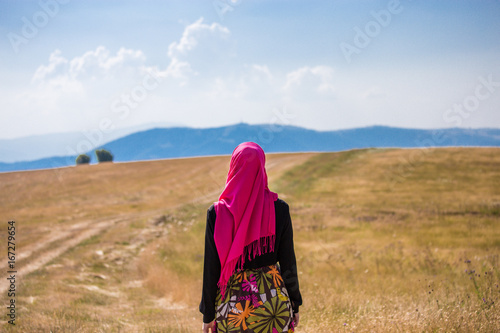 Covered muslim caucasian girl with pink hijab and colorful long skirt standing on a field in summer and looking at the horizon with mountains and blue sky with clouds