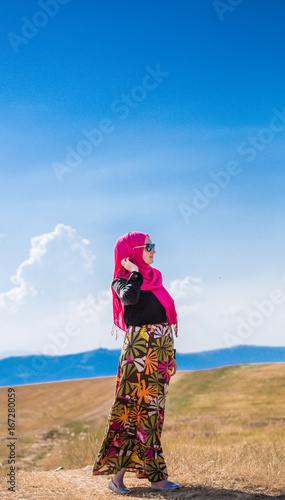 Covered beautiful muslim caucasian girl with pink hijab, colorful long skirt and sunglasses posing from side on grass field in summer with mountains and blue sky with clouds