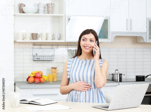 Bild auf Leinwand Young beautiful pregnant woman talking by phone while working in kitchen