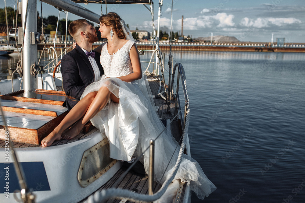 Wedding couple is hugging on a yacht. Beauty bride with groom. Beautiful model girl in white dress. Man in suit. Female and male portrait. Woman with lace veil. Cute lady and guy outdoors